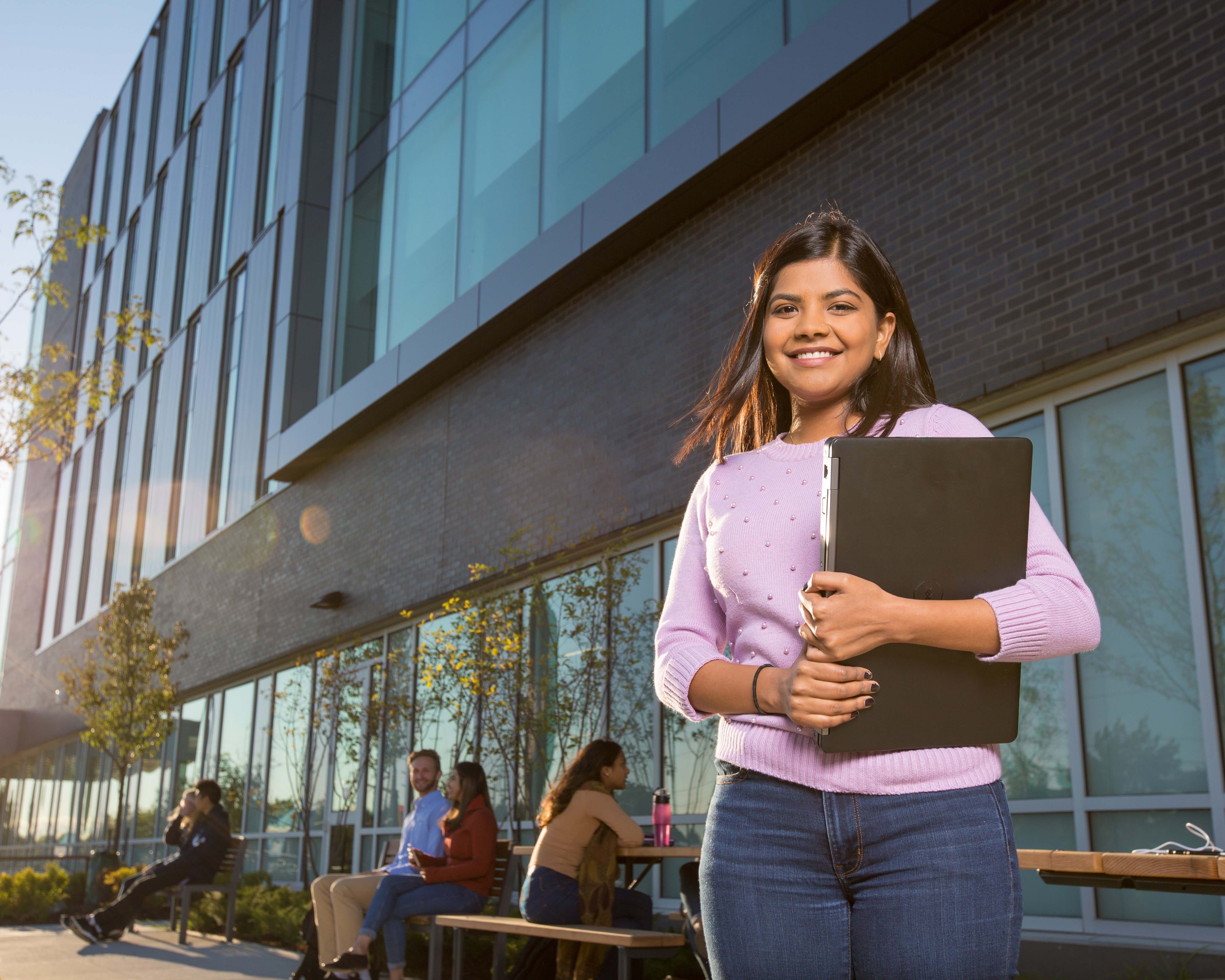 A smiling student stands outside a modern university building, holding a laptop and folder. Other students are seated and chatting in the background, enjoying the sunny day. 